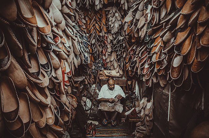 Photograph of Jamal sitting and crafting a shoe in a small room whose walls are covered with hundreds of shoes