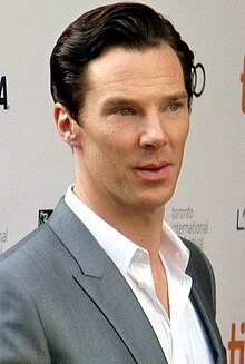 A headshot of a Caucasian male with dark hair; he wears a white shirt with an unbuttoned collar and a grey suit jacket.