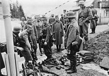 A black and white photograph of German soldiers watching other soldiers laying down their rifles in a pile