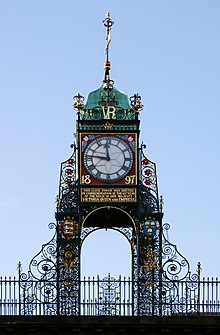 A highly ornate structure in wrought iron, with a railing at the base. It carries a clock with Roman numerals; this has a red frame bearing the date "1897" in gold, and under it is a plate with an inscription in gold lettering. Above the clock are the initials "VR" in gold, and at the summit is a green cupola with a weather vane.