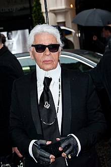 Karl Lagerfeld, who is an older man, wearing a black formal jacket, black gloves, a black tie, and black sunglasses, standing near a Fendi store.