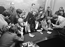 Black and white photo of young white males sat by a long dark desk and surrounded by a crowd of people, including men with microphones attempting to interview them