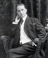 A black-and-white photograph of a seated middle-aged, balding man in a suit and tie, head leaning lightly on his right hand.
