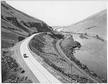 A black and white photograph of a concrete highway curving along a river and railroad in a narrow canyon