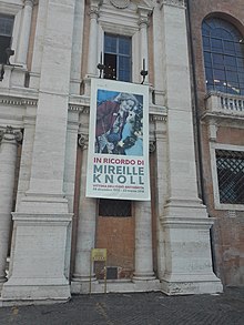 A banner hung off of a balcony in Rome, Italy. It reads "In ricordo di Mireille Knoll, vittima dell'odio antisemita,' along with an image of her smiling and the dates of her birth and death. In English, the banner translates to 'In memory of Mireille Knoll, victim of antisemitism.'