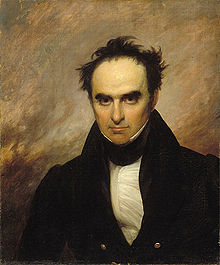 A partially bald black-haired man with large eyes stares directly out of the frame. He is wearing a white shirt, white collar, and black suit.