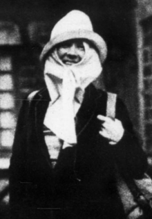 A white woman wearing a brimmed hat, a scarf wrapped around her face, and a dark jacket