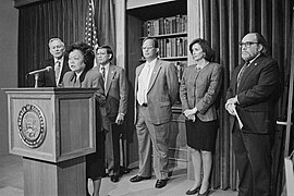 A photograph of a woman standing at a podium, behind whom are a group of four men and one woman standing against a draped wall with a bookcase