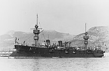 A large, black ship very similar to Suchet, with its bulky masts and short funnels, sits at anchor just offshore.
