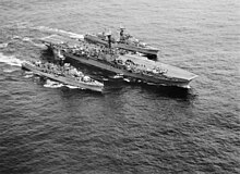 An aircraft carrier sailing in close formation with two destroyers, one on each side.