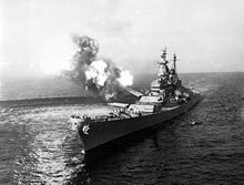 A black-and-white photograph depicting a large gunship sailing toward and slightly to the left of the camera. Guns of various size are visible on the ship, with smoke and flames visible from the turret No. 2 as the gun fires at an unseen target. The pressure from the gun fire has created a disturbance on the water surface.