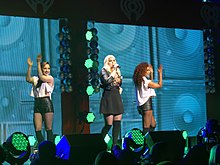 A young long-haired blonde woman singing into a microphone onstage. She sports a black skirt and black Bad Gal jacket. On her left and right are two brunette women dancing, each are wearing a sleeveless white top and leather shorts. A portrait of bass speakers squared in pattern with background colors of neon green as the women's backdrop, as well as the iHeart Radio logo