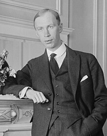 Black and white photo of Prokofiev standing besides a fireplace, his arm rested on the mantlepiece