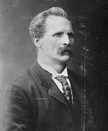 the torso and head of a moustached man, wearing a jacket and tie, viewed from the right