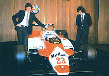 Two men in suits standing left and right of a red-and-white racing car