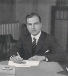 the article subject dressed in a jacket and necktie, writing while seated at a desk