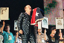 A light-skinned man with brown hair singing into a microphone on a stand, which has a flag draped over it. His shirt and trousers are both grey and feature a design of many overlapping circles. He faces to the right. A line of women stand behind him, each one holding up a sign that says "Donde Estan" or "Justicia". Every sign has an image of a different person below the text.