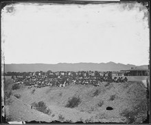 A black and white photograph showing a group of approximately 150 Native Americans in European clothing standing at the slope of a ditch with an arid desert landscape in the background