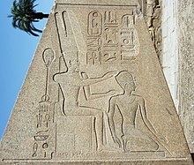 A trapezoidal stone obelisk engraved with Egyptian hieroglyphs and two figures. The figure on the left is seated and wears a tall crown, both hands outstretched to the figure on the right, kneeling, who wears a much smaller crown.