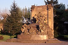 Colour photograph of a sandstone monument in the open air depicting a standing Christ in prayer, surrounded by sleeping disciples and soldiers in arms.