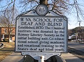 A white historical marker with black lettering illustrating the history of the West Virginia Schools for the Deaf and Blind