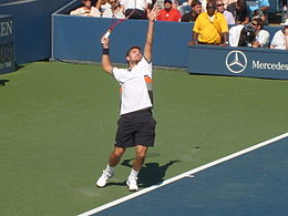 A man wearing a white shirt, black shorts, and white shoes is preparing to serve a tennis ball as he has raised his racket