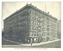 Black and white photo of an urban six-story Victorian commercial building of brick and cast iron on a street corner. The facade to the left is 23 bays wide, to the right is 18, plus a rounded corner one bay wide.