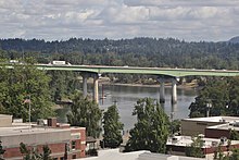 View of a highway bridge on four columns crossing a river with low-rise buildings in the background.