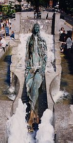 Figure of a young woman sitting on a slope with legs crossed. It is in the middle of a rectangular fountain, surrounded by flowing water.