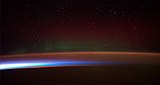 Earth's night-side upper atmosphere appearing from the bottom as bands of afterglow illuminating the troposphere in orange with silhouettes of clouds, and the stratosphere in white and blue. Next the mesosphere (pink area) extends to the orange and faintly green line of the lowest airglow, at about one hundred kilometers at the edge of space and the lower edge of the thermosphere (invisible). Continuing with green and red bands of aurorae streching over several hundred kilometers.