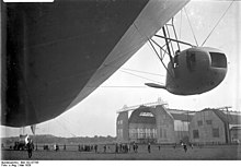 A black-and-white photograph from under the airship's hull while on the ground. In the right middle ground a crewman wearing a leather cap is leaning out of one of the engine nacelles. The wooden grain is visible in the two-bladed propeller, which is stationary and horizontal. The rear engine nacelle is visible and the bottom of the fin. Around 30 people are visible, and about 10 are around the rear nacelle. One man is walking briskly towards the camera. In the background are two large hangars, of unequal size.