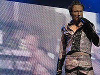 A man in a purple leather suit holds a microphone, standing in front of a large screen.
