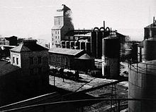 A black-and-white photo of the shale oil processing facility at Kohtla-Järve, dated to 1937. A rail line is shown in the lower third of the photo. A limestone-walled rock hopper tower building and a generator-house are located in the background. Another smaller building is located in the left side of the photo. Several oil tanks are located in the right side of the photo.