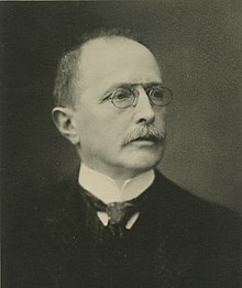 A black-and-white photo of Zuckermandel in a suit and tie wearing glasses