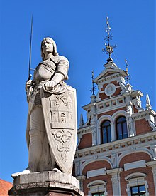 Seitliche Farbfotografie in der Untersicht einer weißen Statue mit Ritterrüstung und Wappenschild. Auf dem Schild befindet sich eine Burg mit Ornamenten. Im Hintergrund ist eine weiß-braune Staffelgiebel mit einem runden Kopfrelief und drei Eisenkreuzen.