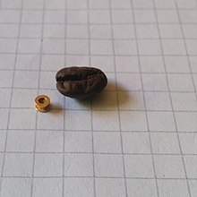 Paukenröhrchen aus Metall, daneben als Größenmaßstab eine Kaffeebohne. Fotografiert auf einem karierten Blatt Papier, ebenfalls als Größenmaßstab (Kantenlänge der Karos 5 mm).