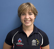Zoe Arancini trägt ein dunkelblaues Hemd und steht vor hinreichend kontrastiertem, hellerem blauen Hintergrund. Auf dem Hemd befinden sich die Logos des Australian Institute of Sport und der Australian Sports Commission.