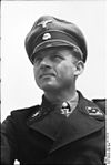 A black-and-white photograph of a man wearing a black military uniform, peaked cap and a neck order in shape of an Iron Cross. His cap has an emblem in shape of a human skull and crossed bones.