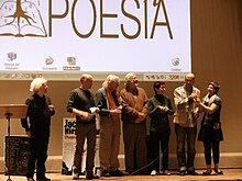 A group of people on stage. They are smiling. There is a projected screen in the background with POESIA written with logo of a book with tree roots coming out of pages. Random alphabets are written on pages.