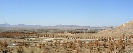 Sparse brown vegetation in dusty soil fills the foreground, fading to distant mountains along the horizon. A barely discernible scattering of buildings is in the middle distance.