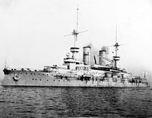 A large warship at rest, with light gray smoke drifting up from its two smokestacks
