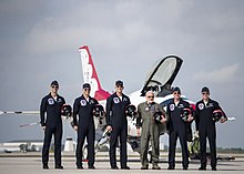 Five men in blue jump suits pose with Aldrin in an olive jump suit on the runway in front of a white F-16