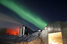 A low shot of a station at the South Pole taken at night. Nearest the front of the photo is a metal structure with a curved roof and a large, open door from which bright light emanates. Slightly further in the distance are two larger buildings. The sky above is a dark blue littered with stars and a green light present across the middle of the sky.