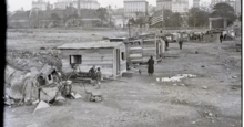 Hooverville with a few people around the wooden shanties, buildings are in the distance