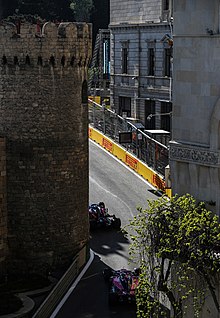 A tarmac race course, lined with concrete and metal barriers, passes between a building and a turret.