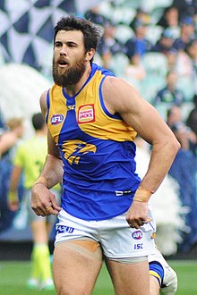 A brown-haired footballer in a sleeveless blue-and-yellow guernsey running on a grassed oval