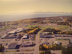 Aerial view of part of downtown Kenai. The intersection of Willow Street and Barnacle Way is in the center of the foreground. Cook Inlet and Mount Redoubt are in the background.