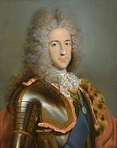 A young man wears a powdered wig while posing in a suit of armour.