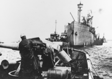 A photograph of two ships. In the foreground is the open bow of USS Rambler with an anti-submarine gun mounted to the left of the frame. USS Bridgeport is in the background of the photo with the bow pointing towards Rambler. There are four smaller ships beside Bridgeport, two on each side.
