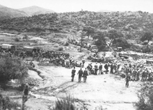 A large group of soldiers, some standing and some sitting, are in a clearing below a hill covered in low grass and a few trees that can be seen in the background. In the middle ground, a line of military vehicles is parked on a road running front-on towards the camera, before hooking to the left in profile. Soldiers work around the vehicles, unloading stores.